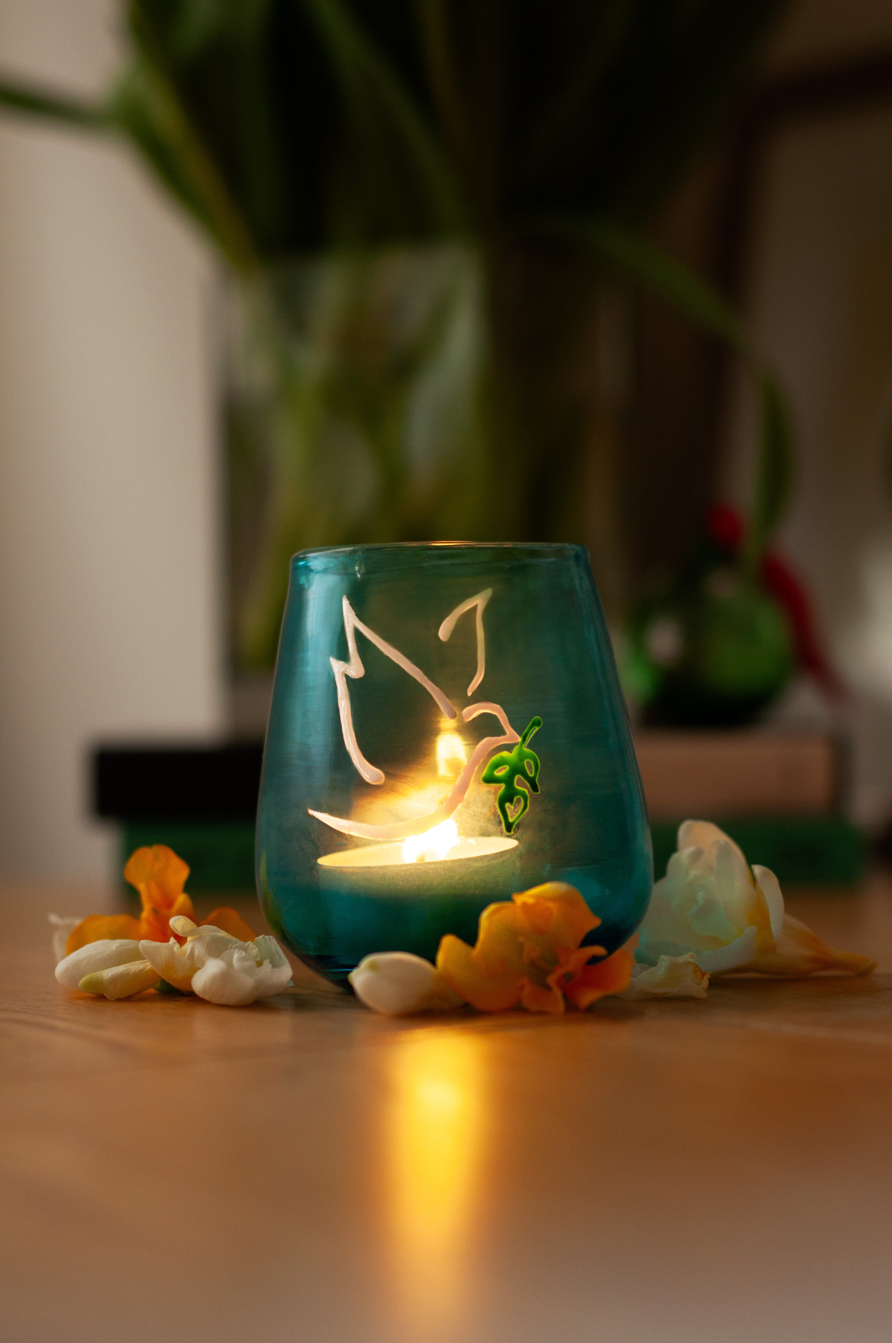 A hand-blown turquoise glass Peace Light with a lit candle inside with painted symbols of a dove and olive branch on the glass surface.  Made in Bethlehem